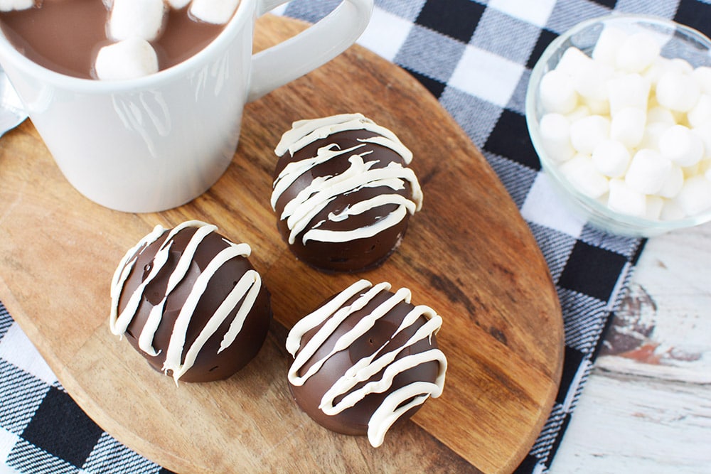 Hot chocolate bombs on a board next to a mug of hot cocoa and marshmallows.