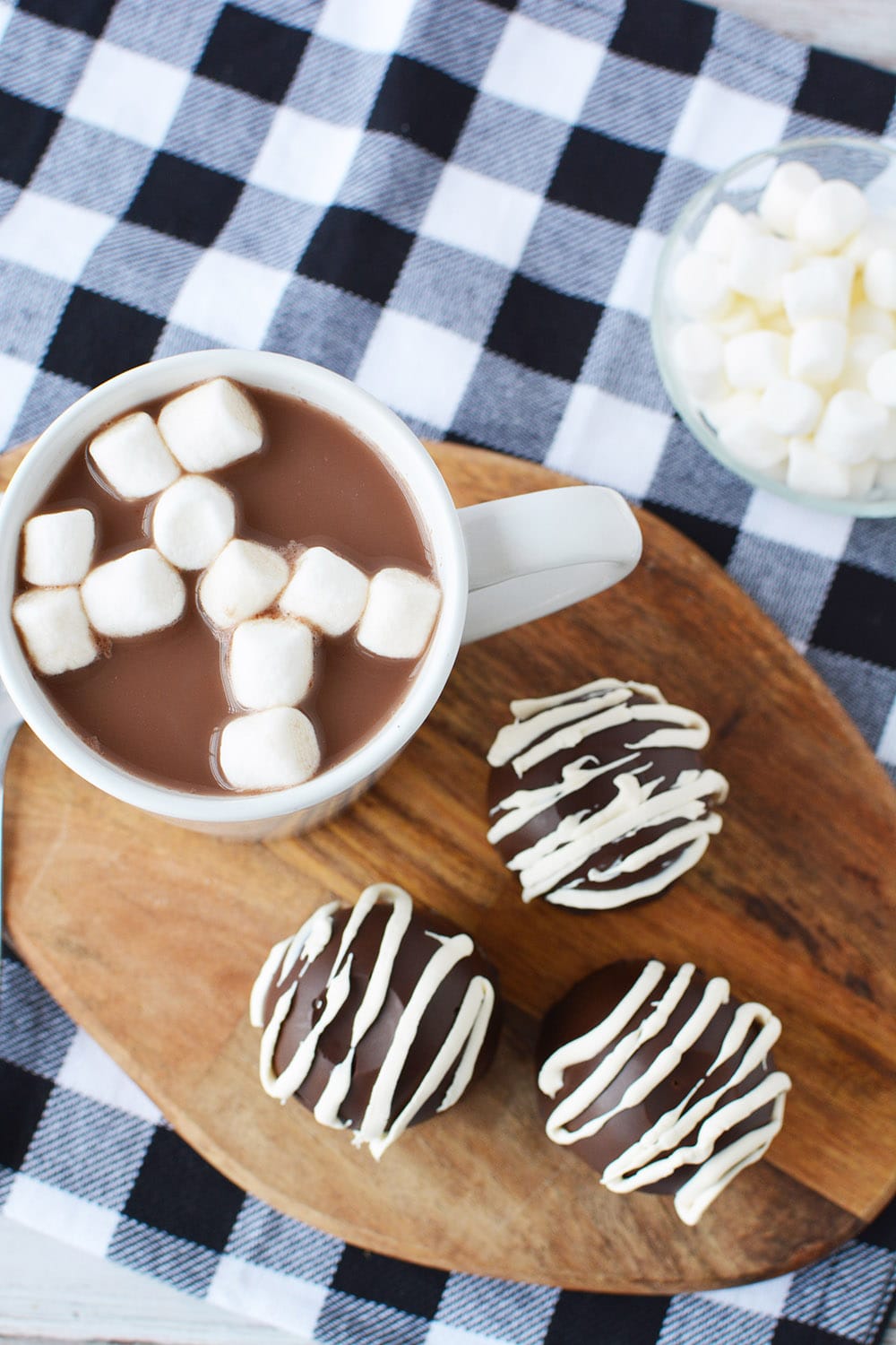 Three hot chocolate bombs next to a mug of hot chocolate with marshmallows in a bowl and a blue checked napkin.