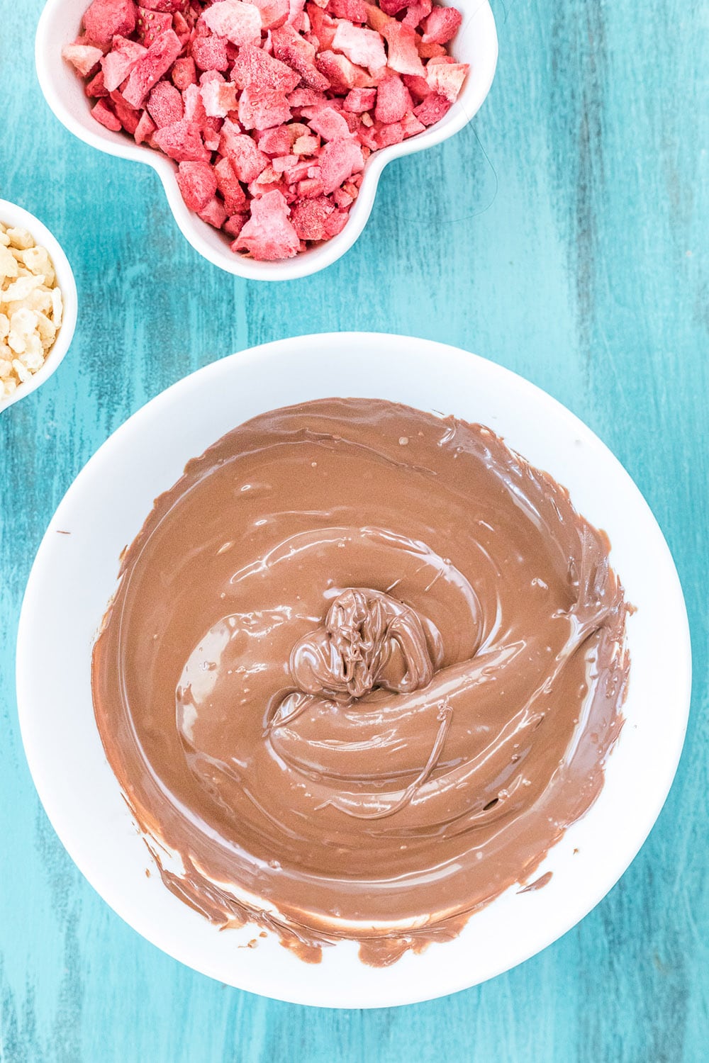 Melted chocolate in a bowl next to strawberries on a blue table.