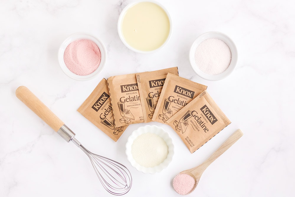 Gelatin packages, whisk, spoon, and bowls of gelatin mix and sweetened condensed milk. 