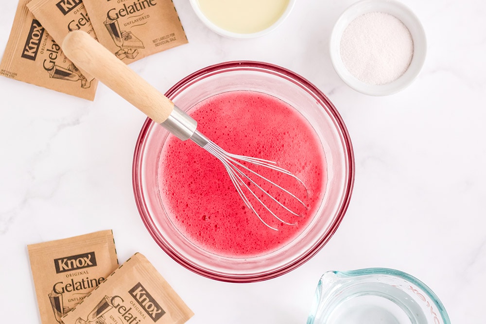 Red gelatin in a bowl with a whisk in it and surrounded by other ingredients on a table.