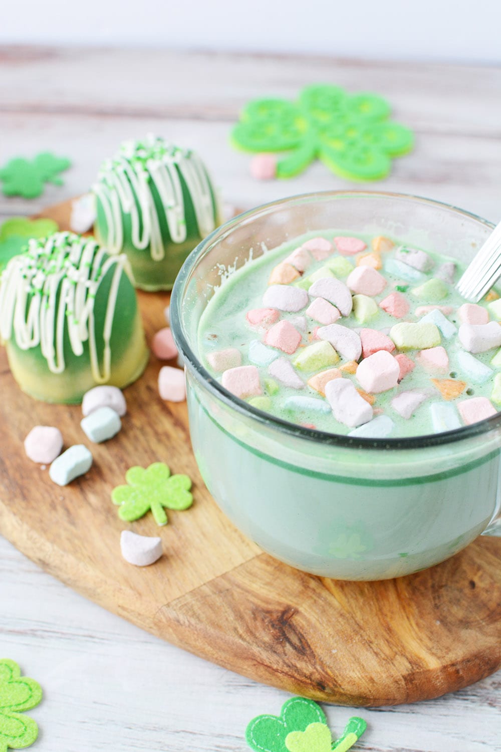 Glass mug of green hot cocoa on a board with cocoa bombs sitting next to it.