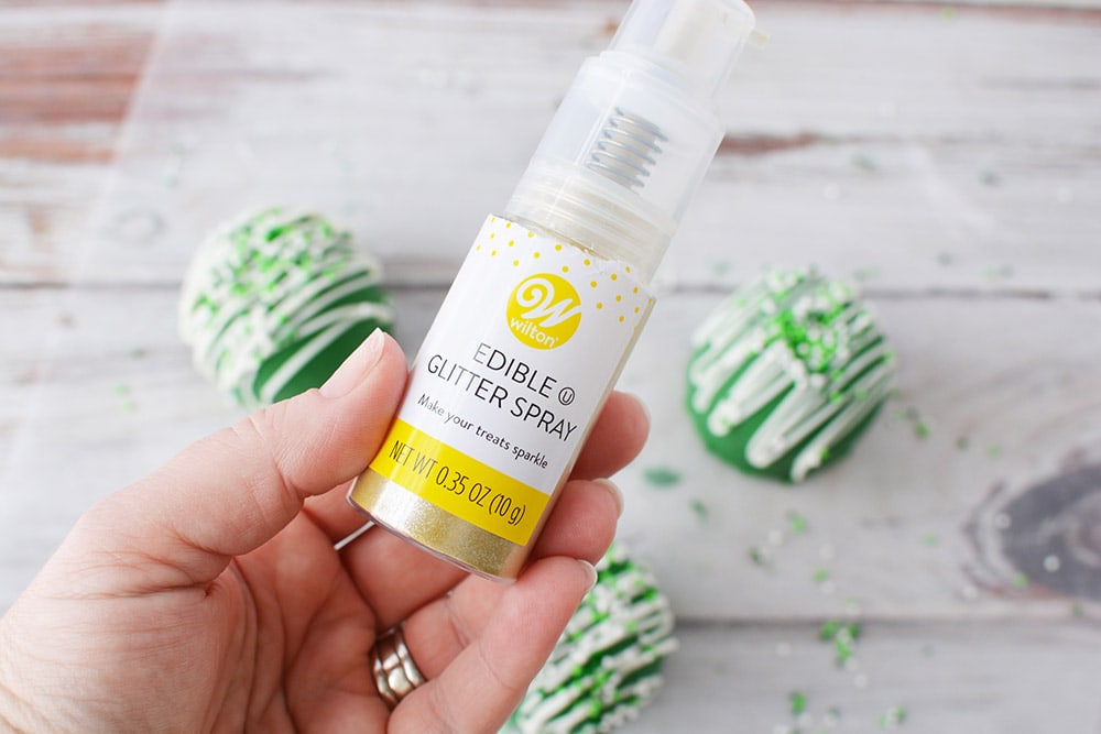 Holding edible gold glitter spray above St. Patrick's Day hot cocoa bombs.