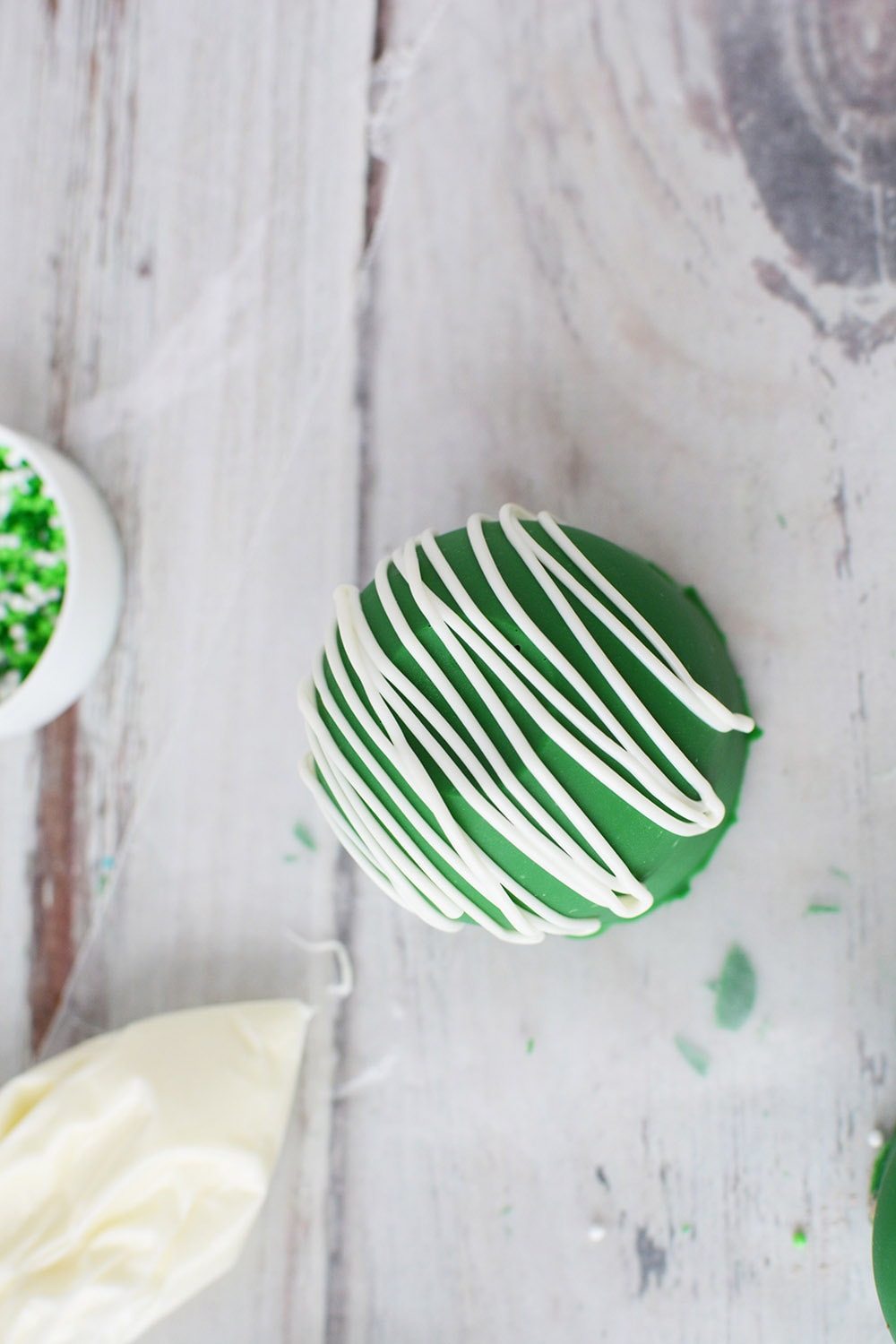 Green cocoa bomb on waxed paper to drizzle white candy topping.