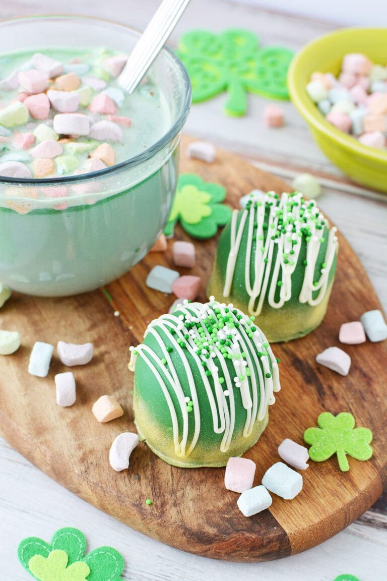 Mug filled with green hot chocolate with rainbow marshmallows next to cocoa bombs and sprinkles.