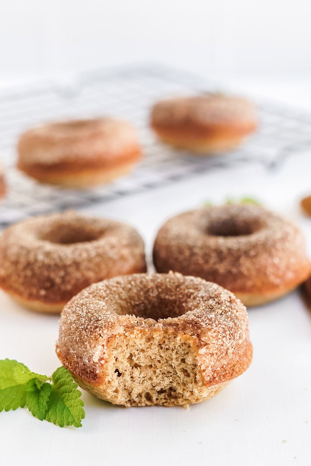 Three apple cider donuts on a table with more on a rack in the background. One donut has a bite taken out of it.