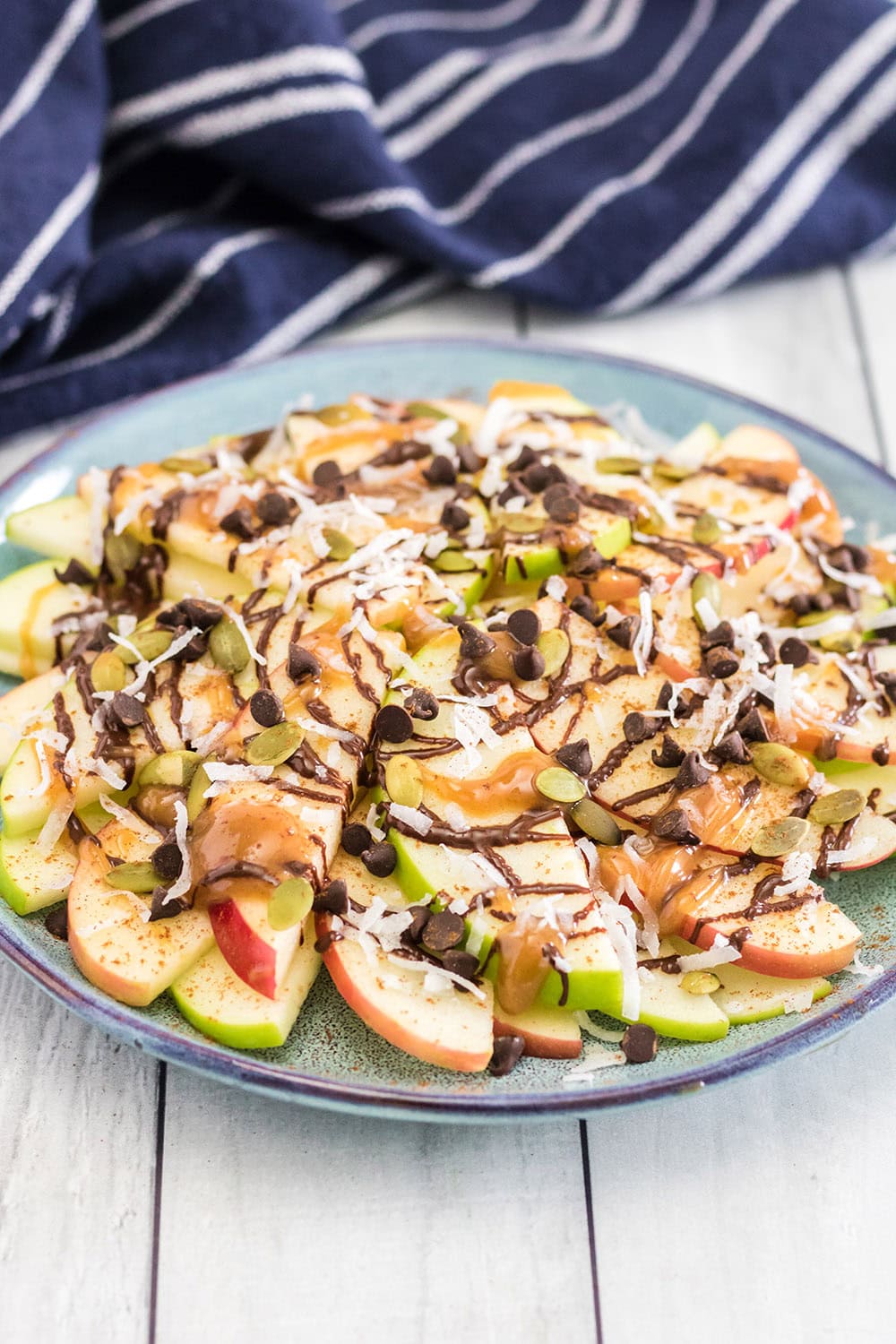 Plate full of apple nachos topped with chocolate, caramel, and nuts with a blue napkin on the table.