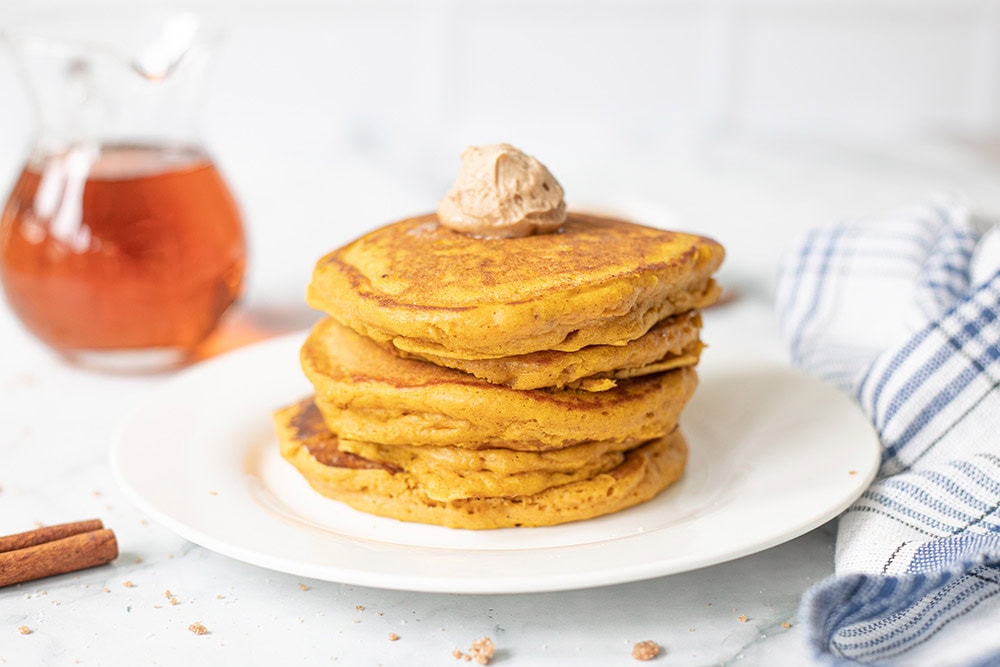 Pancakes stacked on a plate with syrup in the background.