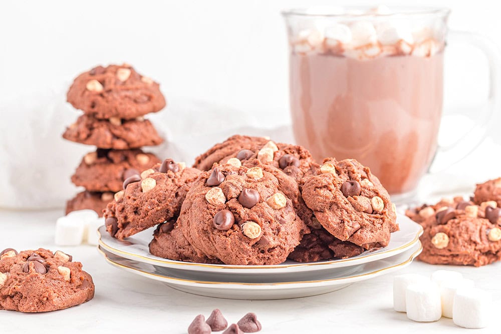 Stacked hot cocoa cookies next to a mug and a plateful of cookies. 
