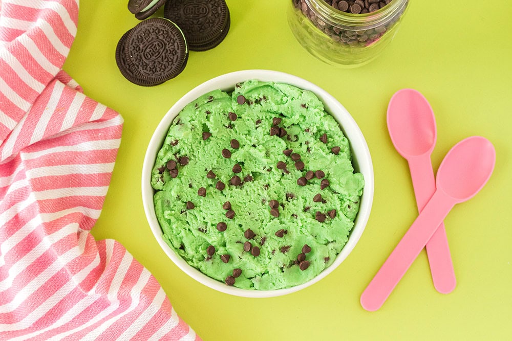 Overhead image of mint cookie dough with chips and pink spoons on a green table.