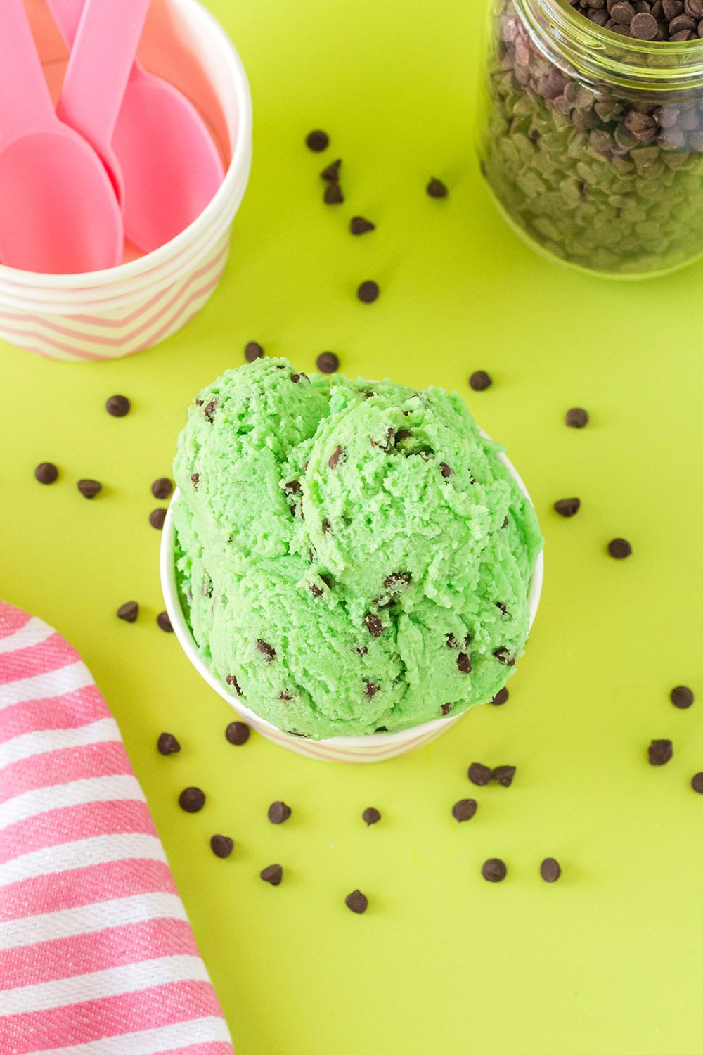 Overhead shot of mint chip cookie dough on a green table surrounded by mini chips and pink spoons.