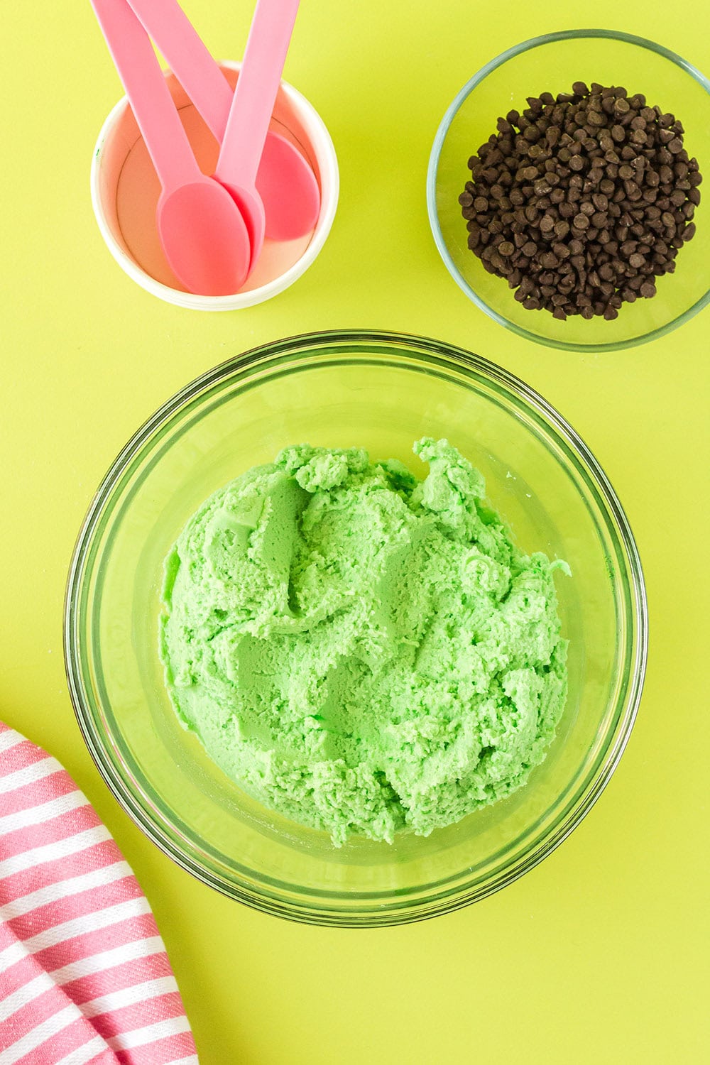 Green cookie dough in a mixing bowl next to chips and pink spoons.