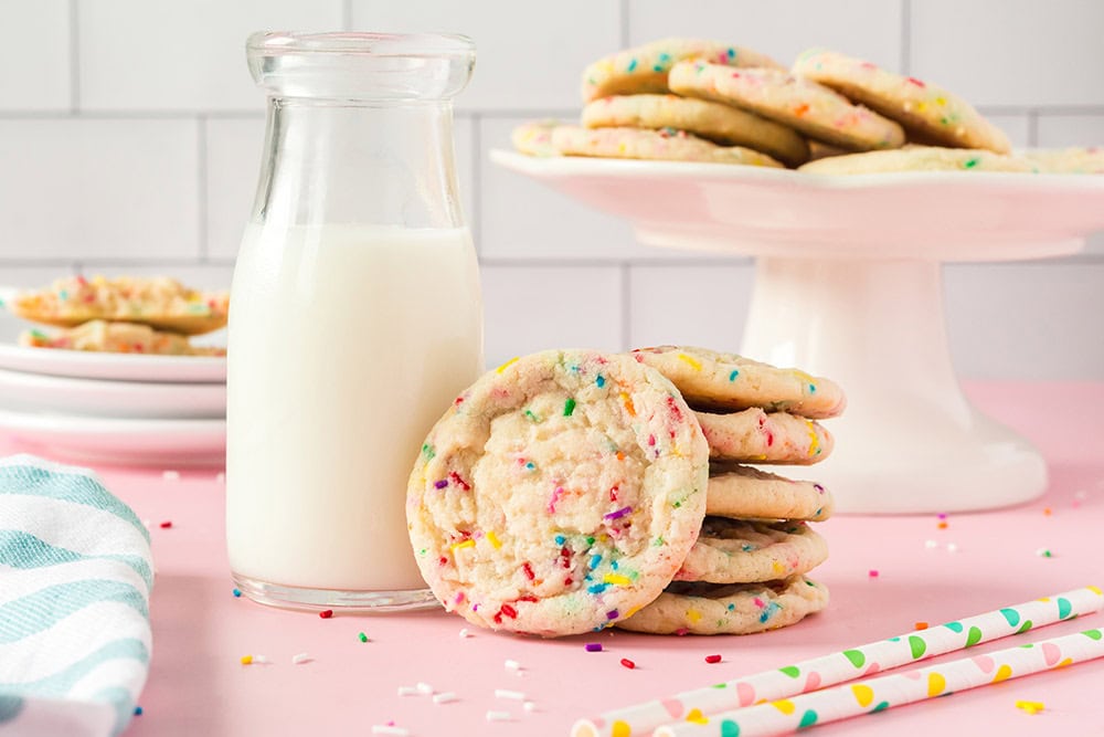 Funfetti cookies on a stand, stacked in front of the stand, and next to a glass of milk. 