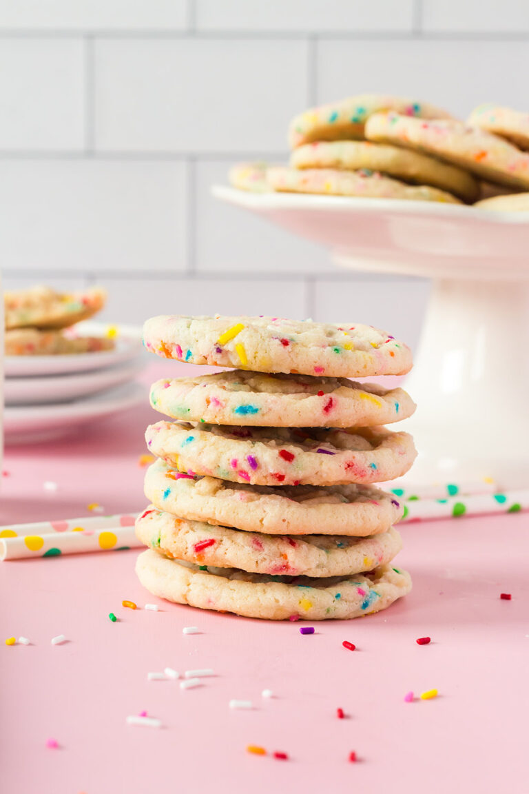 Stack of sugar cookies with rainbow sprinkles next to cookies on a stand and plates.