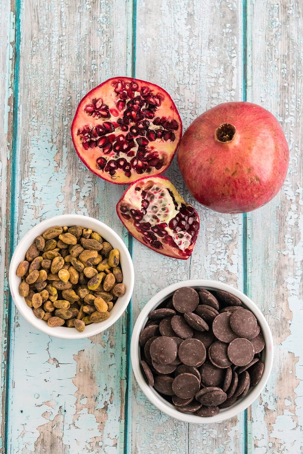 A sliced pomegranate, bowl of pistachios, and bowl of dark chocolate wafers on a table.