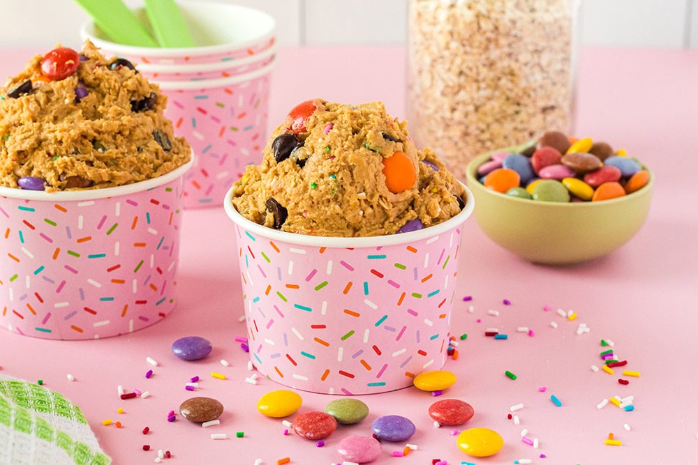 Cups of monster cookie dough next to M&M candies and oats. 