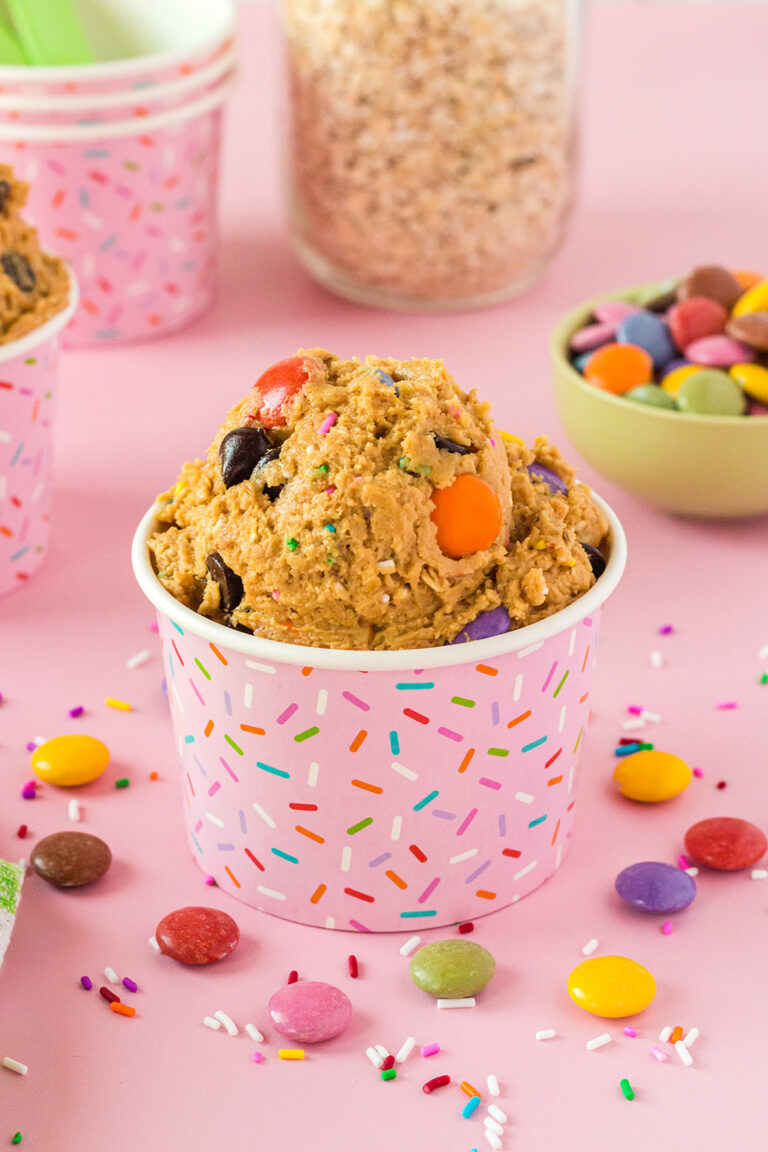 Pink cup with monster cookie dough in it and candy pieces on a pink table.
