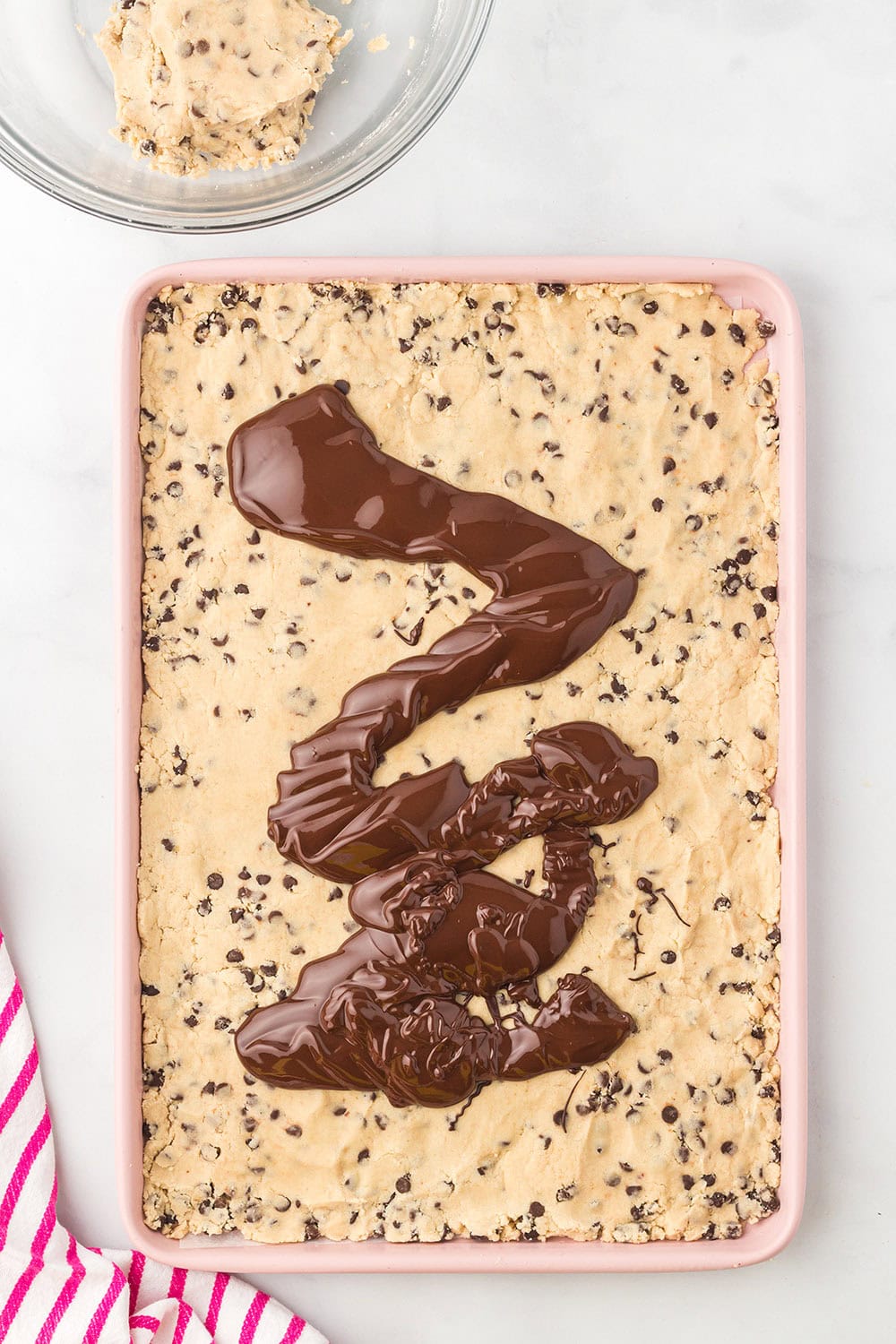 Pouring melted chocolate on top of cookie dough in a baking sheet.