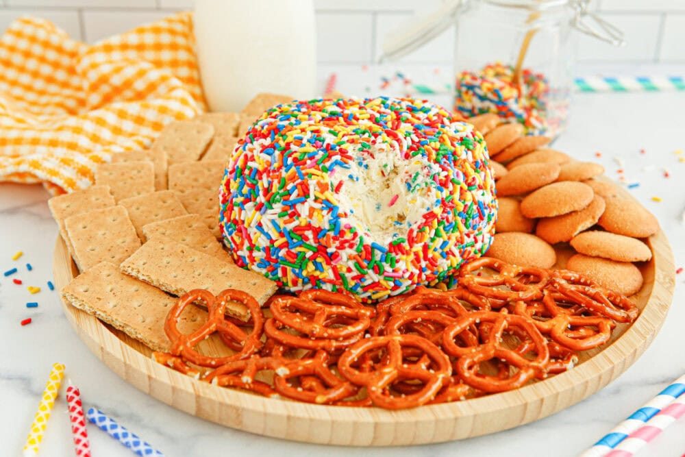 Cream cheese ball covered in sprinkles on a plate with pretzels, graham crackers, and cookies. 