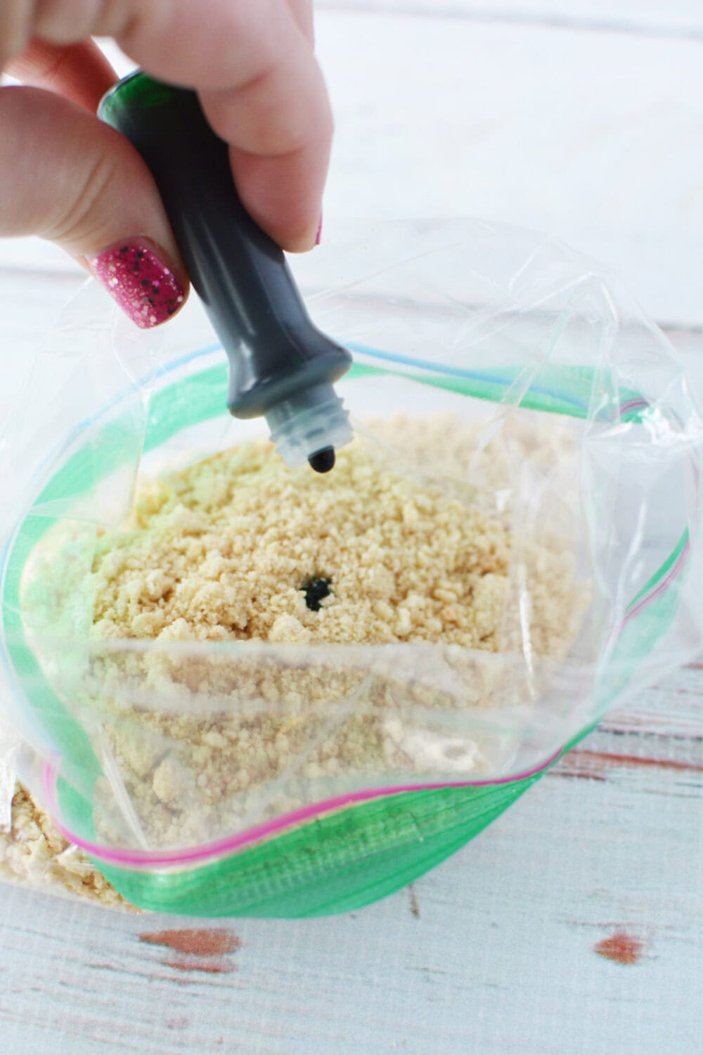 a hand holding green food coloring over a bag of crushed cookies