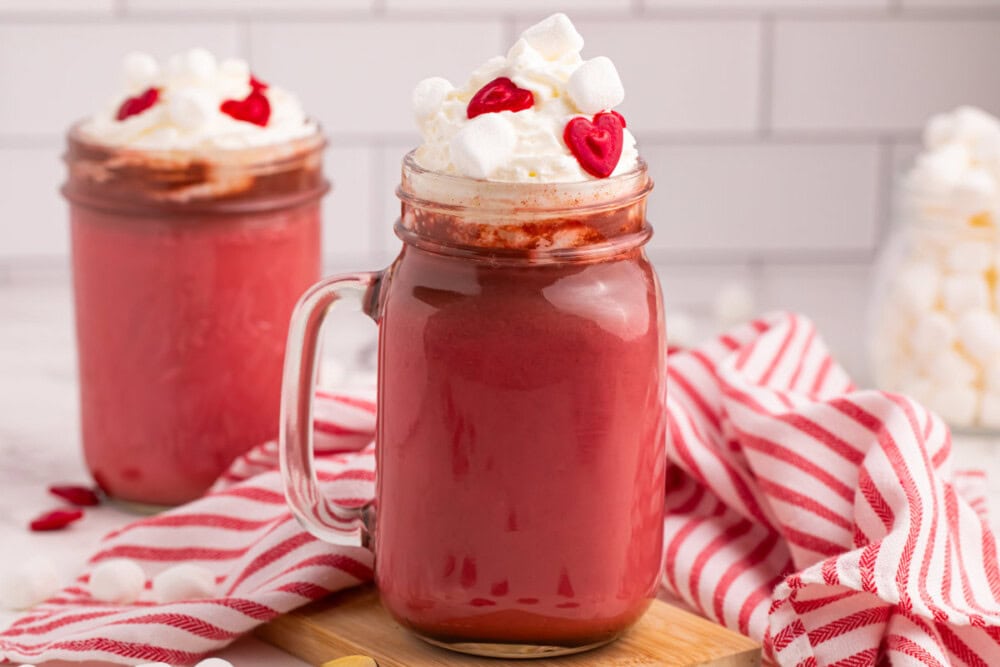 Glass jar filled with red velvet hot chocolate topped with whipped cream and heart sprinkles.